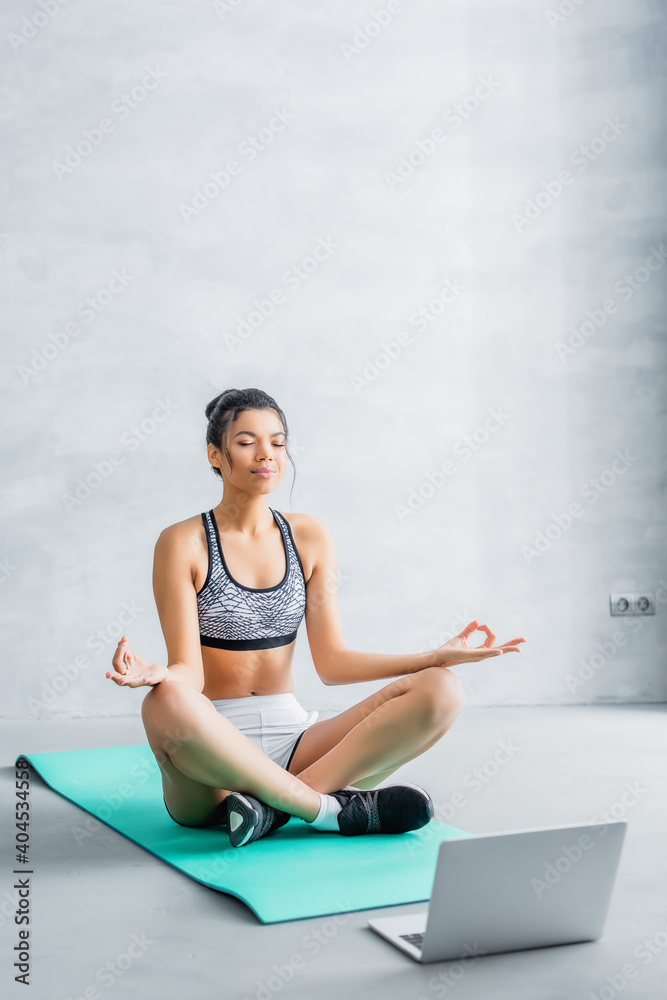 Fototapeta premium young african american woman in sportswear meditating in lotus pose on fitness mat near laptop