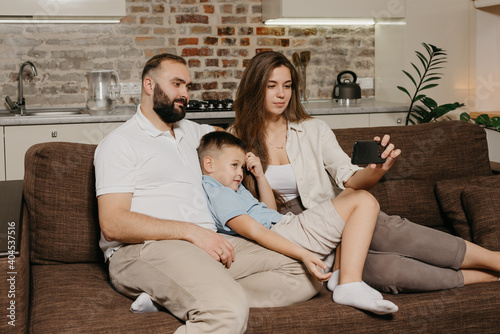 A dad, a son, and a young mother are watching a video on a smartphone on the sofa. A mom with long hair is demonstrating to her family news on the screen of the cellphone in the evening at home.