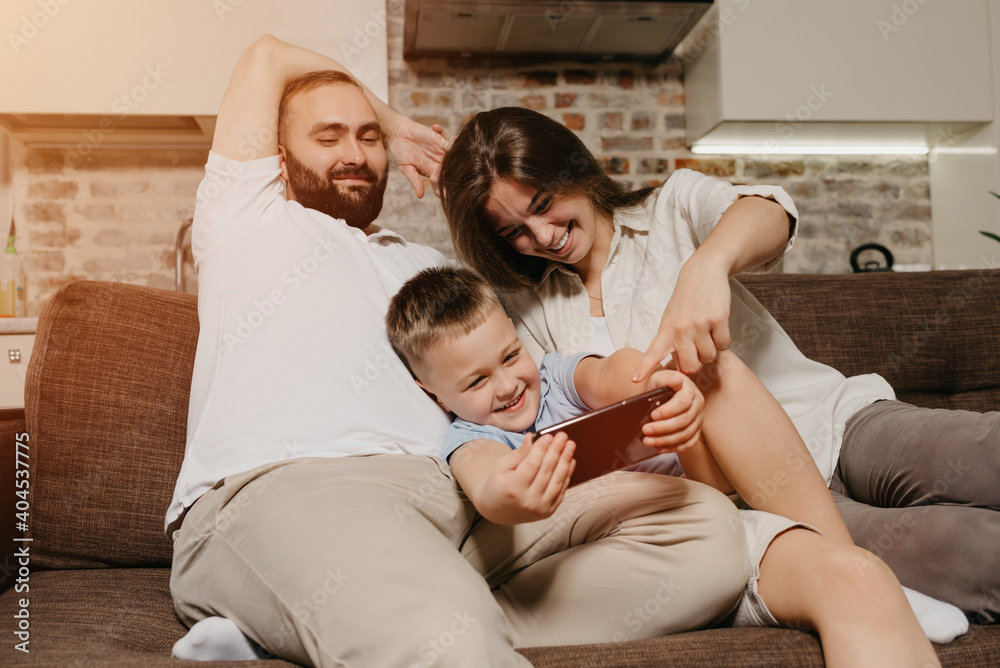 A dad, a son, and a mom on the sofa. A happy child is enjoying the game on the smartphone near smiling mother and father in the evening at cozy home. A dad, a son, and a mom on the sofa. A happy child is enjoying the game on the smartphone near smiling mother and father in the evening at cozy home.