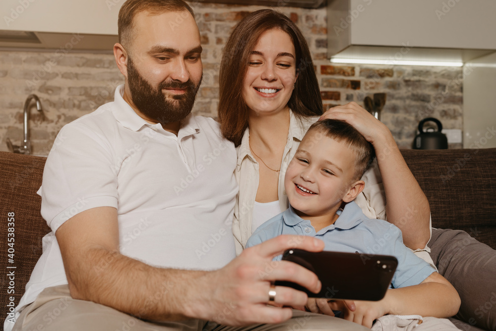 Obraz premium A dad, a son, and a mom are watching video on the sofa. A happy husband with a beard is demonstrating the show on the smartphone to a smiling child and a wife in the evening at home.