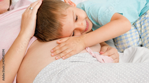 Portrait of cute smiling toddler boy hugging and stroking his pregnant mother with big belly. Concept of loving children and family happiness expecting baby