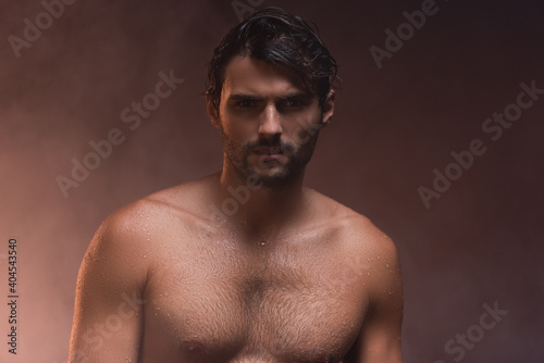 wet, shirtless man looking at camera while posing on dark background with smoke