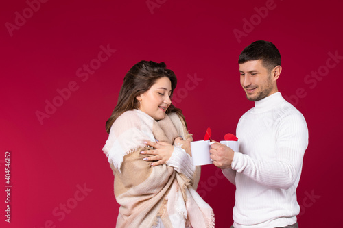 A girl wrapped in a plaid drinks warm tea with her beloved man on a red background with an empty side space.