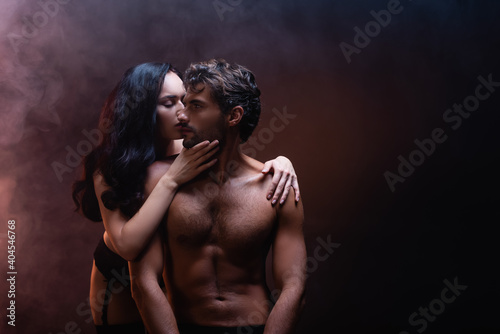 seductive woman embracing and kissing sexy shirtless man on dark background with smoke