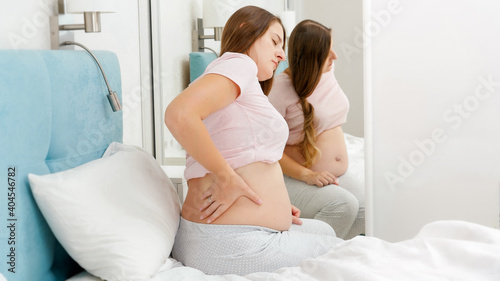 Beautiful pregnant woman in pajamas touching and massaging her aching back. Concept of pregnancy healthcare and medical examination.