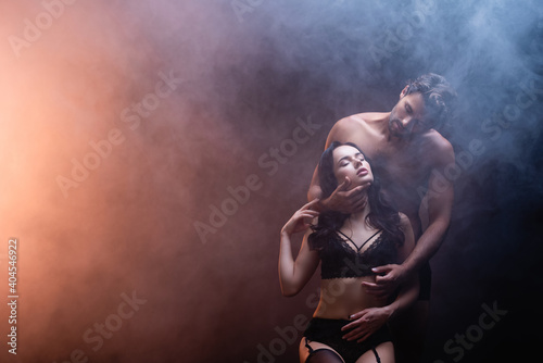 shirtless muscular man embracing neck of sexy woman in black lace underwear on dark background with smoke