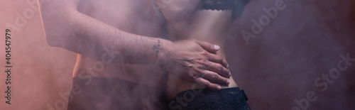 cropped view of shirtless tattooed man embracing woman on dark background with smoke, banner