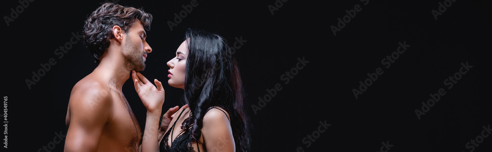 side view of seductive woman touching chin of sexy muscular man isolated on black, banner side view of seductive woman touching chin of sexy muscular man isolated on black, banner