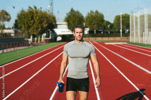 Mature fitness man on red running track, walking and looking at blue bottle in hand. Front view. Copy space. Lifestyle and sport