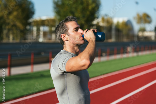 Mature fitness man on red running track, drinking from blue bottle. Copy space. Close up. Lifestyle and sports