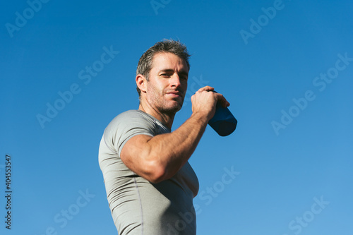 Mature fitness man looking up with blue sky background, holding blue bottle in hand. Copy space. Close up. Lifestyle and sports