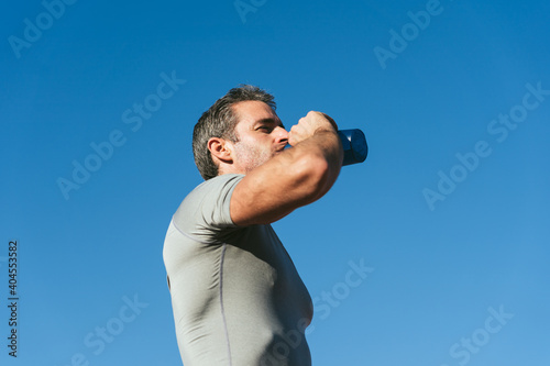 Mature fitness man with blue sky background, drinking from blue bottle. Copy space. Close up. Lifestyle and sports photo