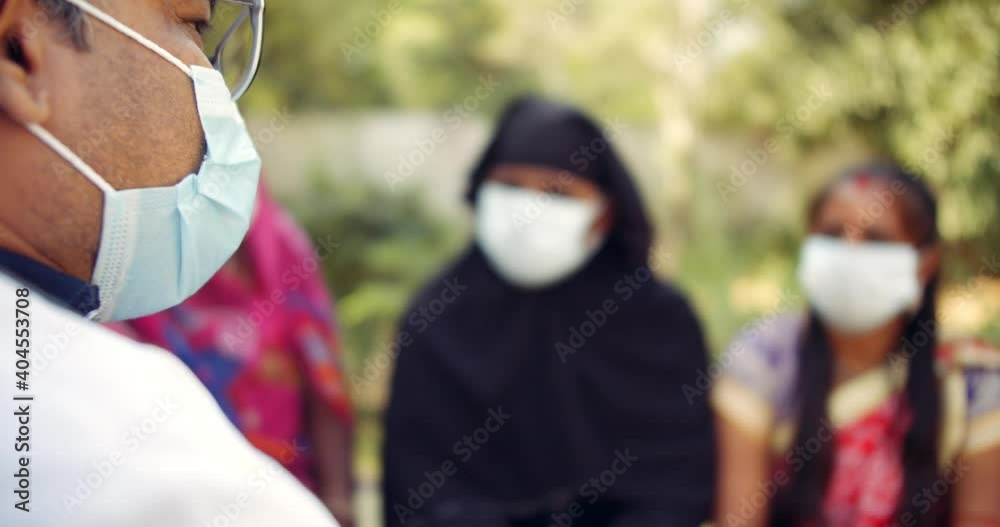 Shallow focus bokeh of close-up face of a doctor wearing face mask outdoors in nature public park informs a group of people wearing colorful vibrant traditional dress about coronavirus covid-19 Shallow focus bokeh of close-up face of a doctor wearing face mask outdoors in nature public park informs a group of people wearing colorful vibrant traditional dress about coronavirus covid-19
