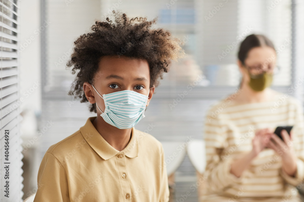 Portrait of teenage African-American boy wearing mask and looking at camera while waiting in line at medical clinic, copy space