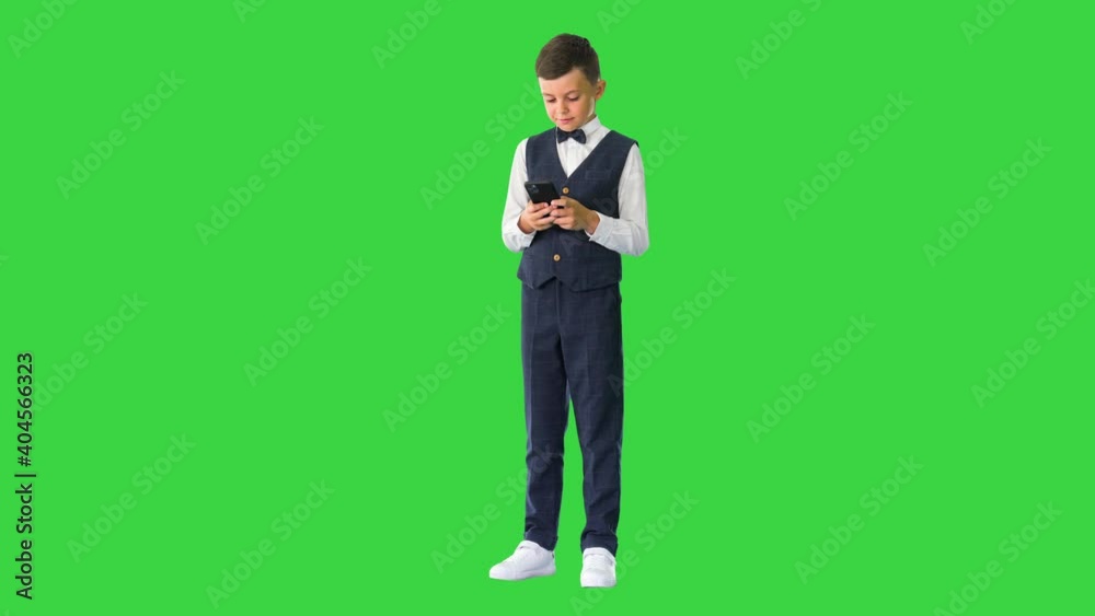 Smiling boy in a white shirt, bow tie and vest texting on the phone on a Green Screen, Chroma Key. Smiling boy in a white shirt, bow tie and vest texting on the phone on a Green Screen, Chroma Key.