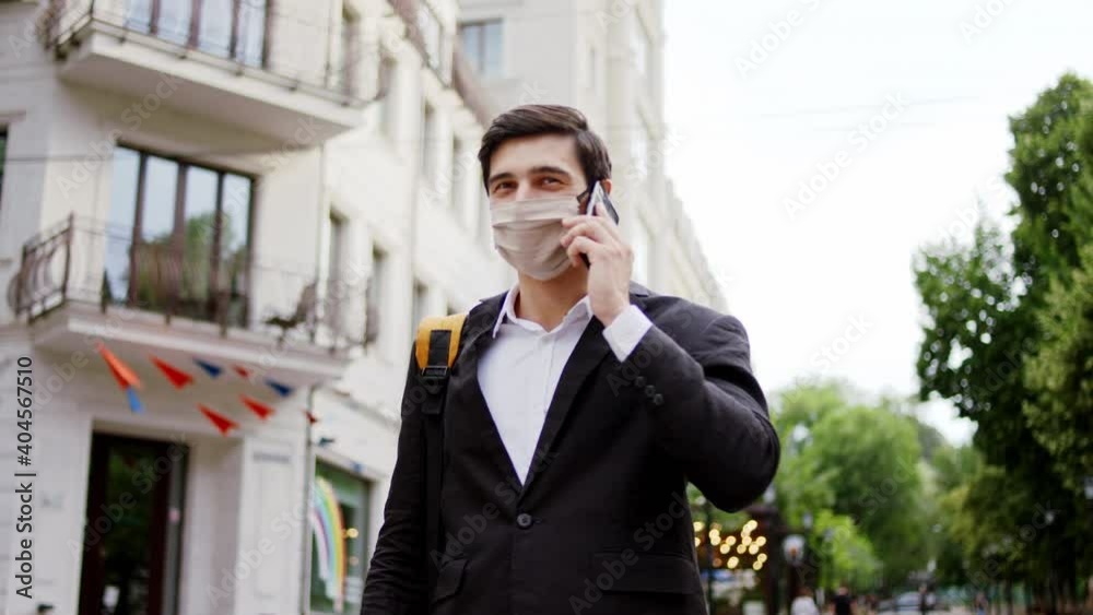 Charismatic businessman with a protective mask walking down on the street and speaking on his smartphone in front of the camera Charismatic businessman with a protective mask walking down on the street and speaking on his smartphone in front of the camera