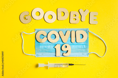 Words GOODBYE COVID 19 from wooden letters, syringe with vaccine on yellow background, and facemask. Vaccination and the end of epidemic concept.