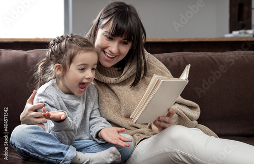 Young mother with her daughter are reading a book at home while sitting on the couch.