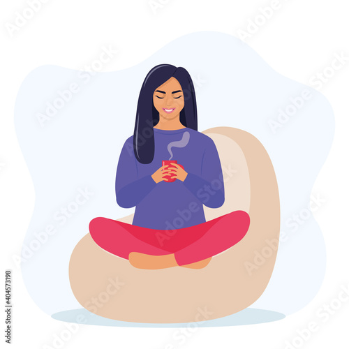 Young woman sitting on bean bag in a Lotus pose and holds a mug of hot drink in her hands. Cozy rest and relaxation concept. Vector illustration.