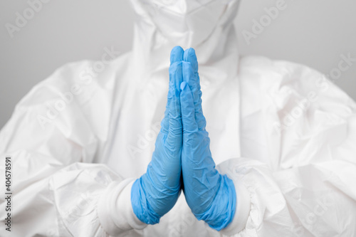 Doctor in a biohazard protective PPE suit and protective face mask against coronavirus, Sars-Cov-2, Covid-19 praying in blue rubber gloves.
