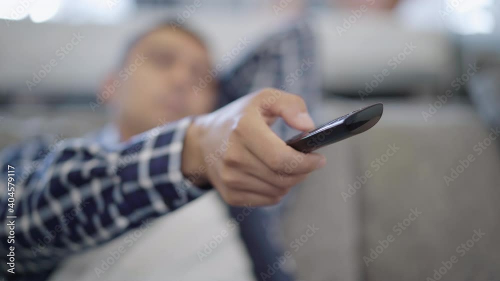 Close-up of remote control in male Caucasian hand switching channels with blurred young man lying on couch at background. Unrecognizable relaxed guy resting at home on weekends alone. Close-up of remote control in male Caucasian hand switching channels with blurred young man lying on couch at background. Unrecognizable relaxed guy resting at home on weekends alone.