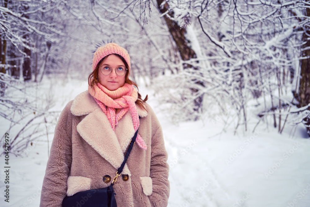 A woman in a white fur coat and pink hat stands in a snow-covered winter forest. Portrait of an adult woman with glasses and a bag among the trees in the park.