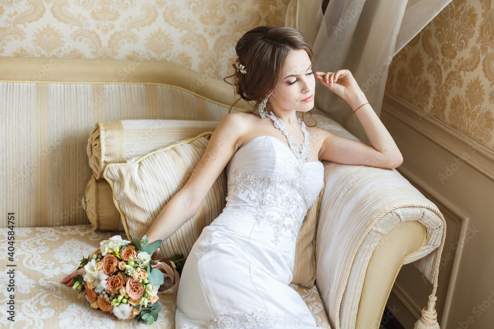 Beautiful young Bride with classic hairstyle sits on a sofa in a bedroom. Classic fish-silhouette white wedding dress. Full height portrait 