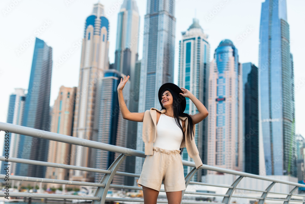 Cheerful traveler woman wave hello walking on a promenade in Dubai Marina district. Travel destinations and tourist lifestyle in UAE