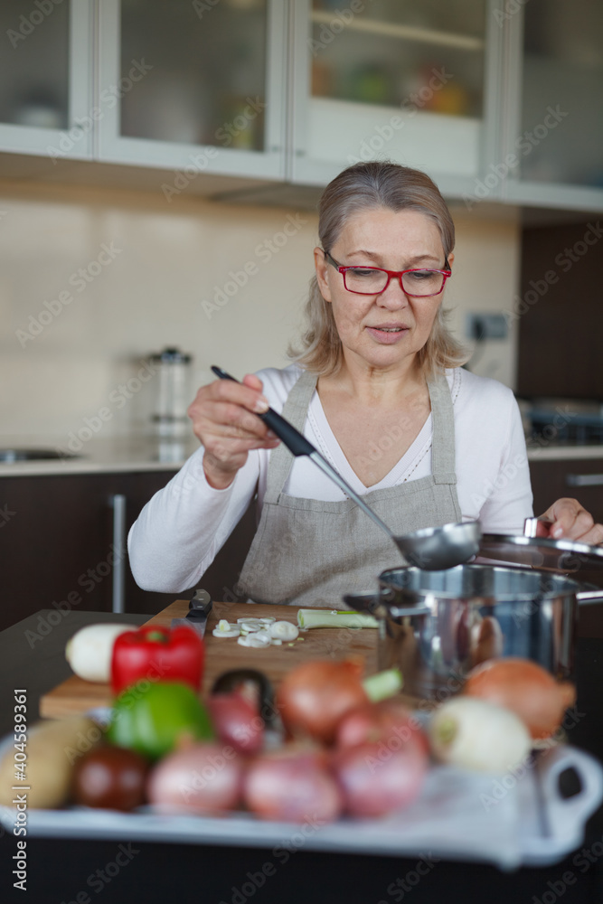 A housewife tastes broth in her kitchen. On the table in front of her are vegetables for the soup.