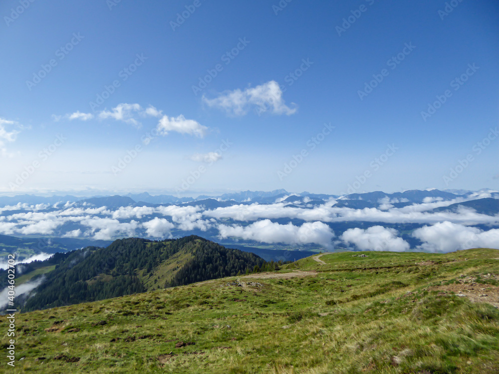 A panoramic view on the Alpine valley from the top of Granattor in Austrian Alps. The valley is shrouded in fog. The slopes are lush green. Many mountain poking above the clouds. Mystery mood