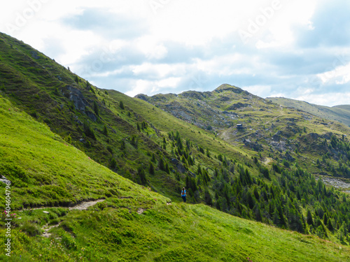 A view on lush green slopes of Austrian Alps in the region of Millstatt. There is a narrow pathway along the slope. Endless mountain chains in the back. Dense forest at the foothill. A bit of overcast