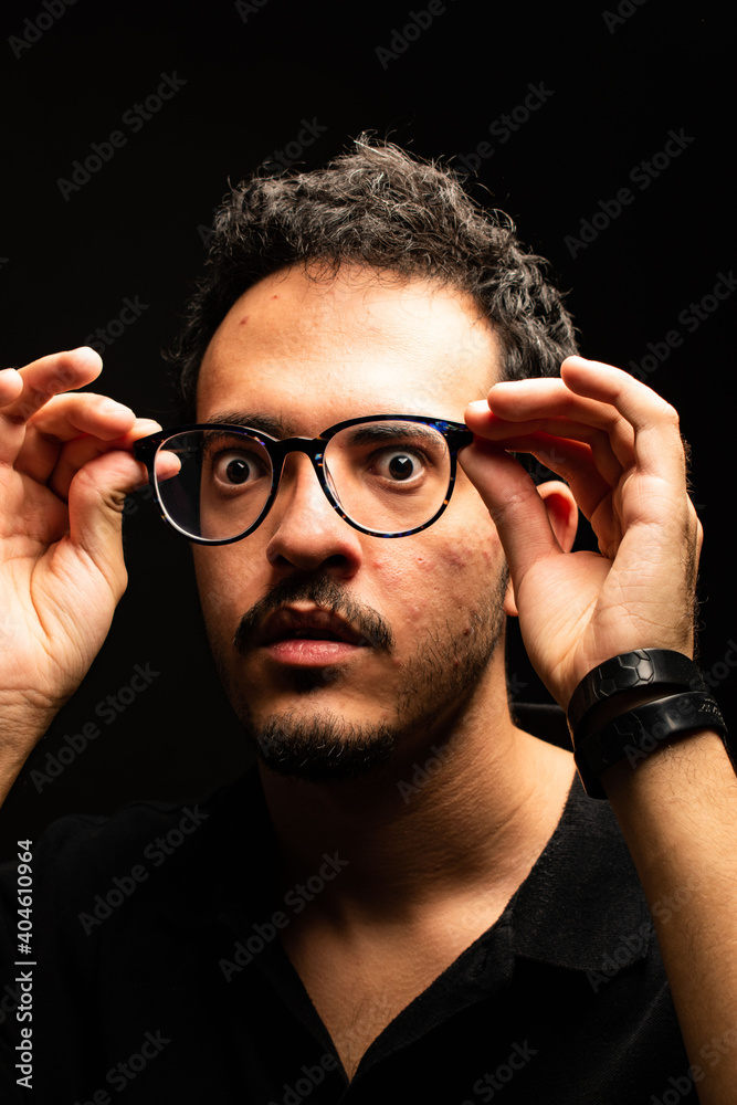 Man with glasses stares at camera dressed in a black polo shirt while holding his glasses away from his face. Black background. Man with glasses stares at camera dressed in a black polo shirt while holding his glasses away from his face. Black background.