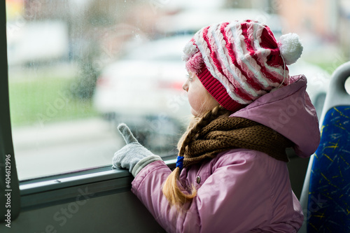 A little girl in a jacket and a hat sits on the bus and looks out the window, pointing at something with her finger.