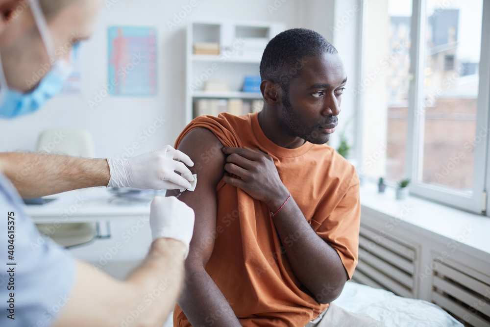 Portrait of adult African-American man looking away while getting covid vaccine in clinic or hospital, with male nurse injecting vaccine into shoulder Portrait of adult African-American man looking away while getting covid vaccine in clinic or hospital, with male nurse injecting vaccine into shoulder