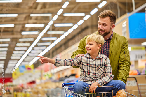 kid boy point finger at side in store during shopping with father, child sits on cart havng fun, enjoying time in grocery supermarket photo