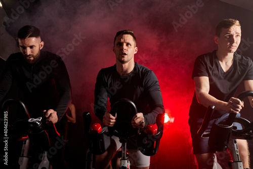 three men riding stationary bikes together during a cardio workout session at the gym isolated in smoky red neon lighted space photo