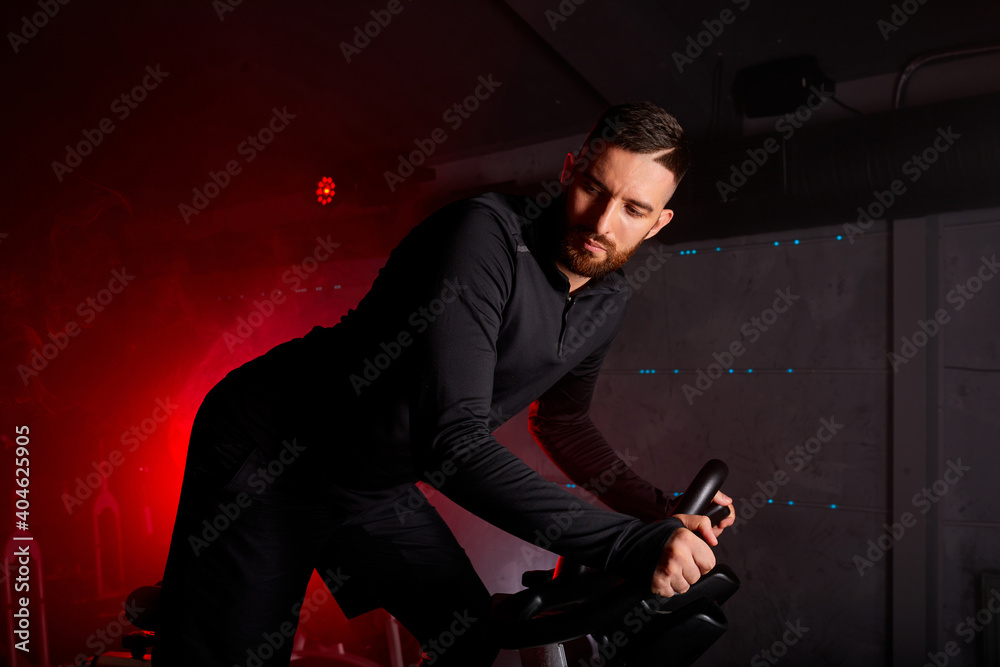 male is engaged in an exercise bike, personal training in the red neon lighted gym, wearing tracksuit male is engaged in an exercise bike, personal training in the red neon lighted gym, wearing tracksuit