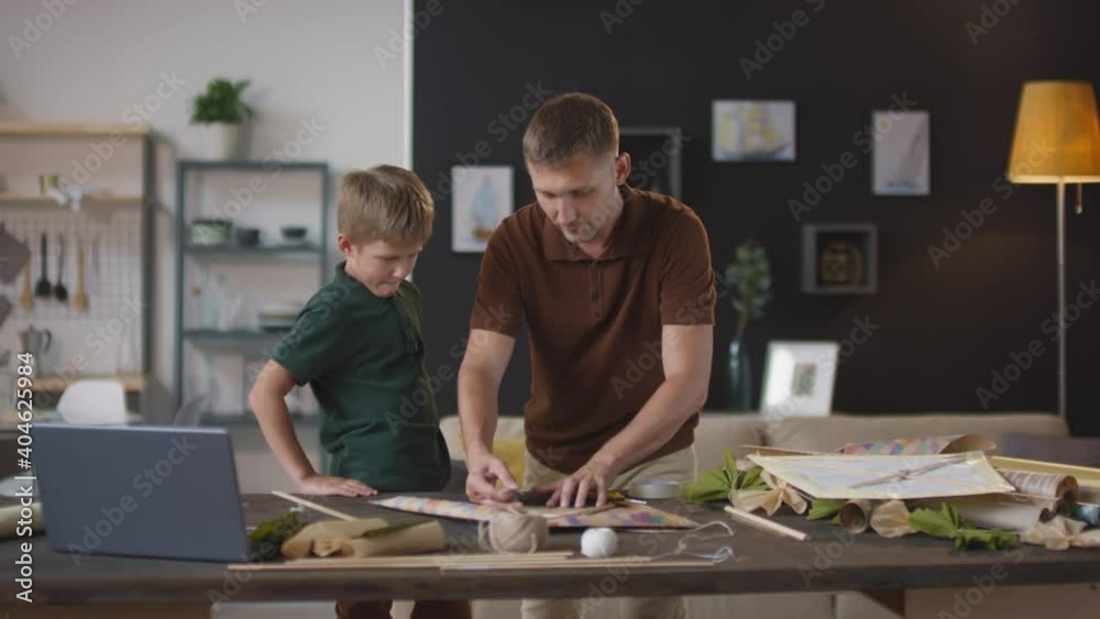 Medium handheld shot of adult caucasian man constructing colorful diy flying kite at home together with son watching process Medium handheld shot of adult caucasian man constructing colorful diy flying kite at home together with son watching process