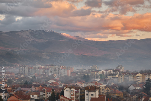Misty, smoky cityscape of Pirot during sunset with impressive, rocky peak lighten by golden light in the background and a cloudy sky