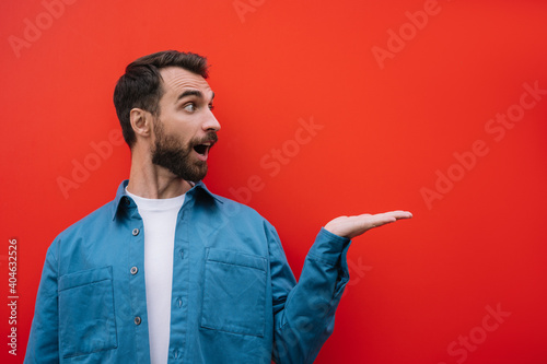 Emotional bearded man wearing casual shirt pointing with hand, isolated on red background. Copy space, advertising concept