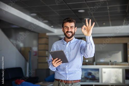 Smiling bearded CEO standing in lobby of export firm, showing okay gesture and holding tablet. Delivery is always on time.