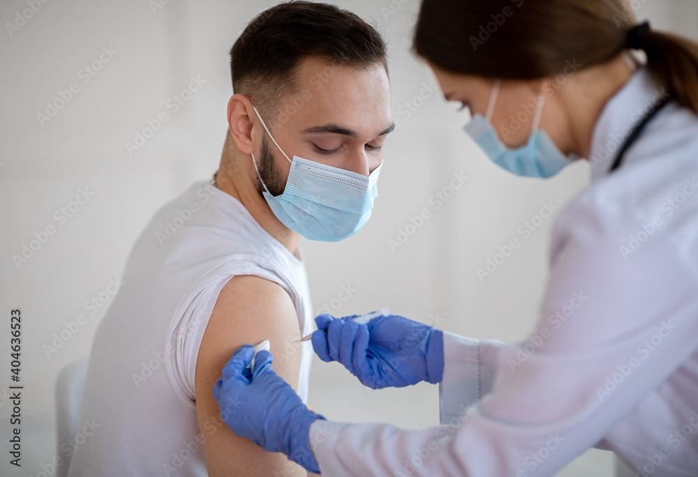 Female doctor making injection of coronavirus vaccine to young patient in medical mask at health centre Female doctor making injection of coronavirus vaccine to young patient in medical mask at health centre