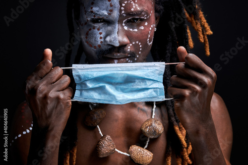 aborigen male wearing medical mask on face during coronavirus, covid-19 all over the world. man with shamanic paintings on face looks at camera isolated on black background photo