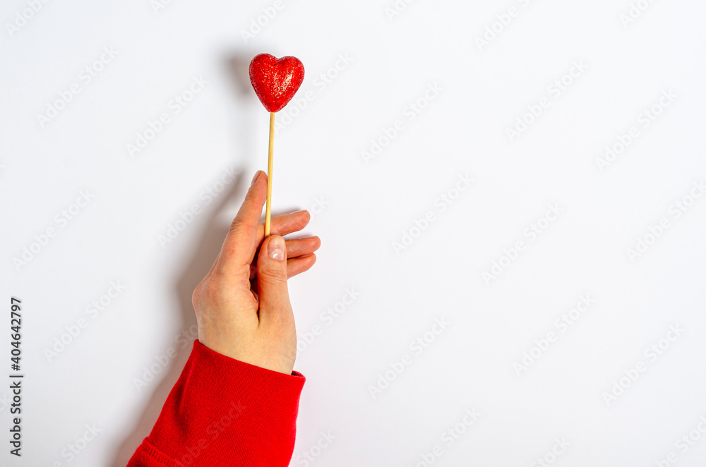 A female hand holds a shiny heart on a stick on a white background.