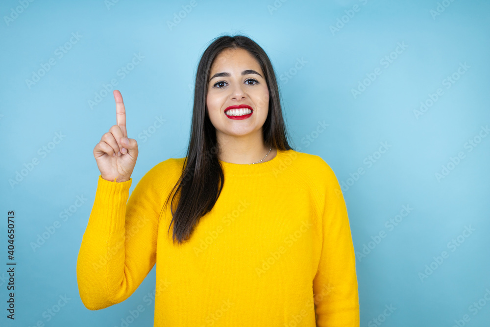 Young beautiful woman wearing yellow sweater over isolated blue background showing and pointing up with fingers number one while smiling confident and happy Young beautiful woman wearing yellow sweater over isolated blue background showing and pointing up with fingers number one while smiling confident and happy