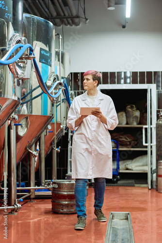 Young female brewery expert in whitecoat entering data in tablet in plant