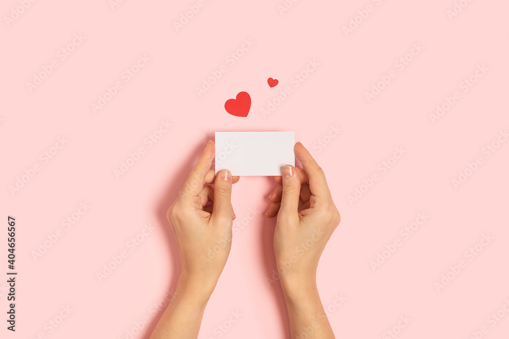 Top view of woman hands holding blank paper greeting or invitation card over pink pastel background with confetti, and heart shape. Christmas, Valentine, New Year, winter holidays concept Top view of woman hands holding blank paper greeting or invitation card over pink pastel background with confetti, and heart shape. Christmas, Valentine, New Year, winter holidays concept
