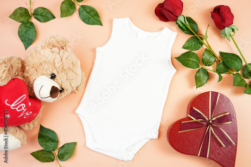 Happy Valentine's Day baby apparel flatlay owith teddy bear, red roses and heart shaped gift on modern coral background. Mock up with negative copy space for your text or design here. photo