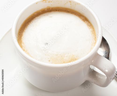 a white cup of coffee with creamy froth on the top on a saucer stands on a white table.