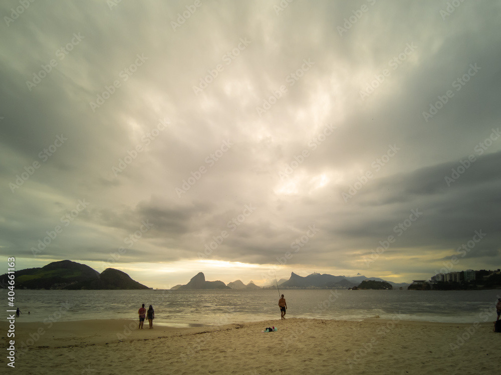 Beach before the storm. Sky with many tropical rain clouds and few people in the distance. Mountains of Rio de Janeiro city on the horizon.